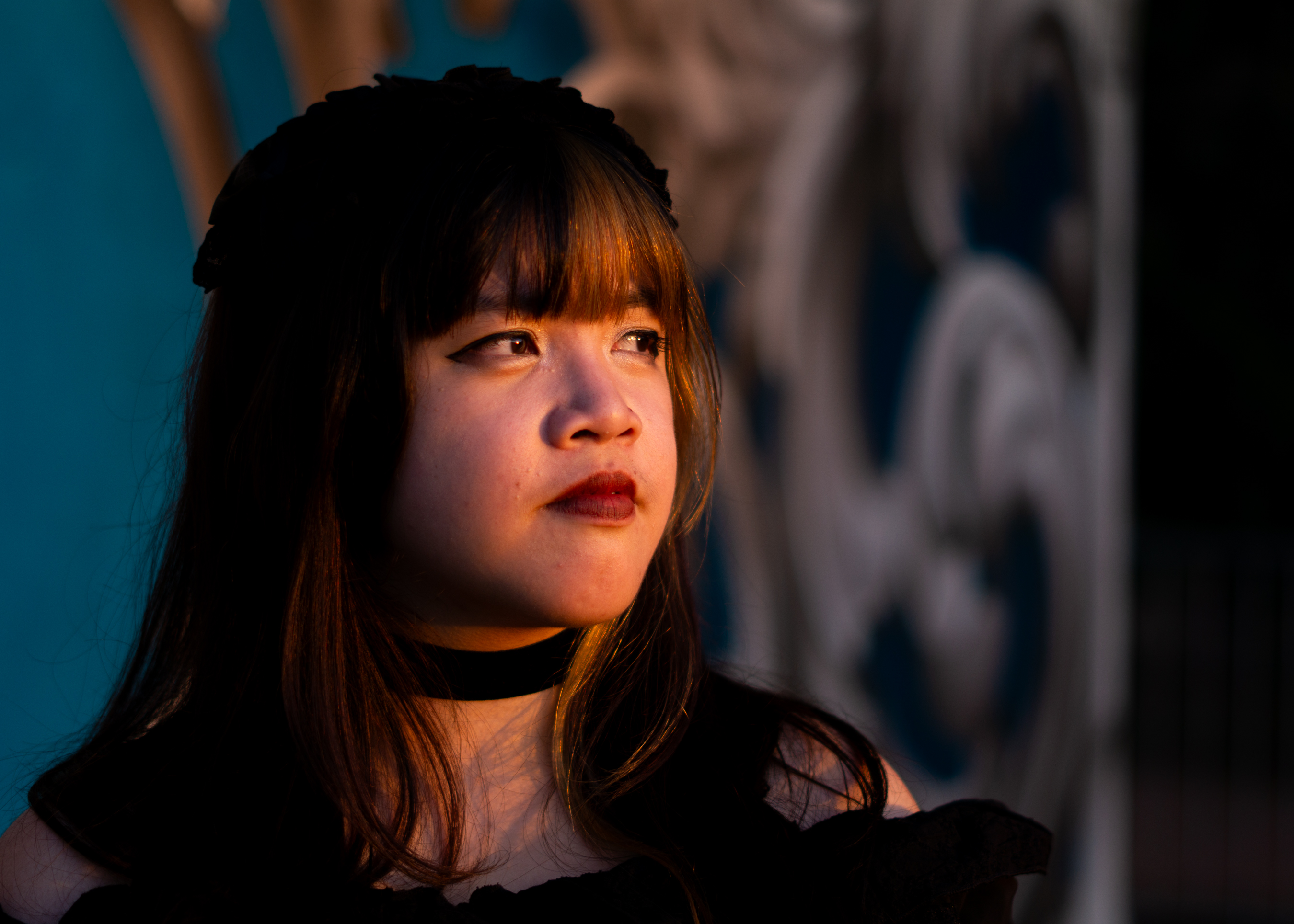 A fem-presenting person in a frilly black dress standing in front of an illustrated wall, blue with white fleurs-de-lys. Their face is illuminated with sunlight as they look to the side.