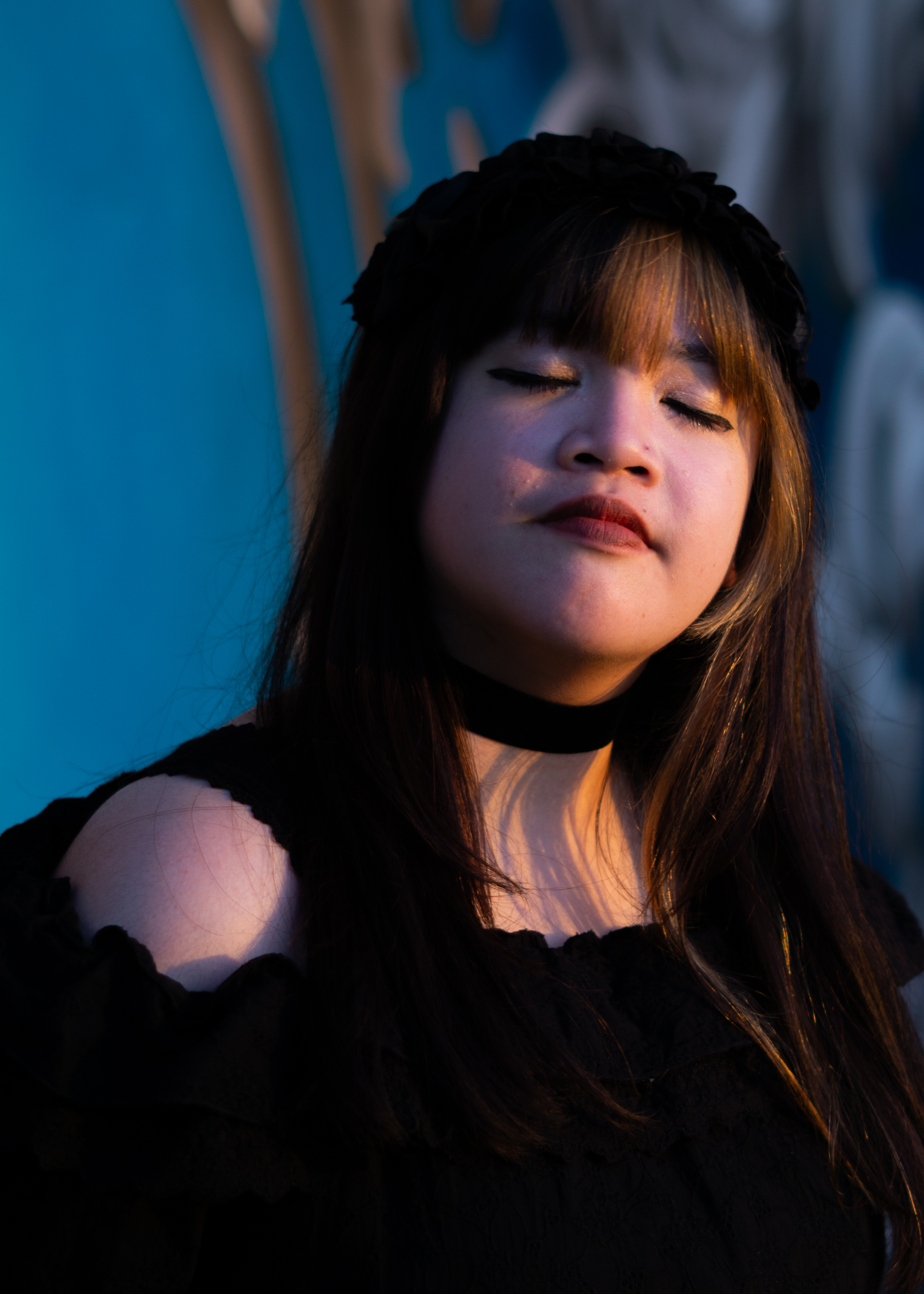 A fem-presenting person in a frilly black dress standing in front of a brick wall, standing in front of an illustrated wall, blue with white fleurs-de-lys. Their eyes are closed and they're tilting their head.