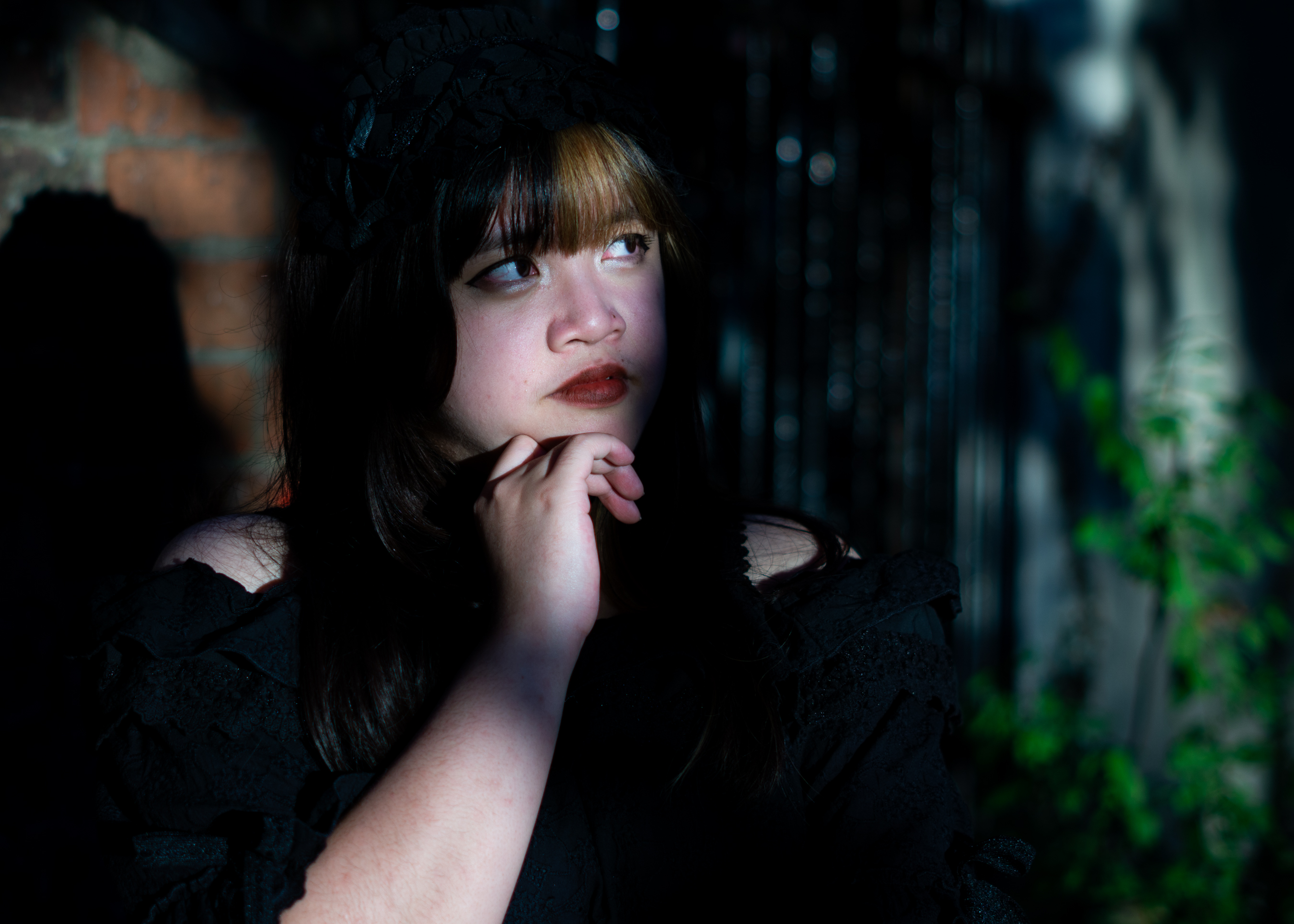 A fem-presenting person in a frilly black dress standing in front of a brick wall, covered mostly in shadow with a streak of sunlight illuminating a stretch of their face.