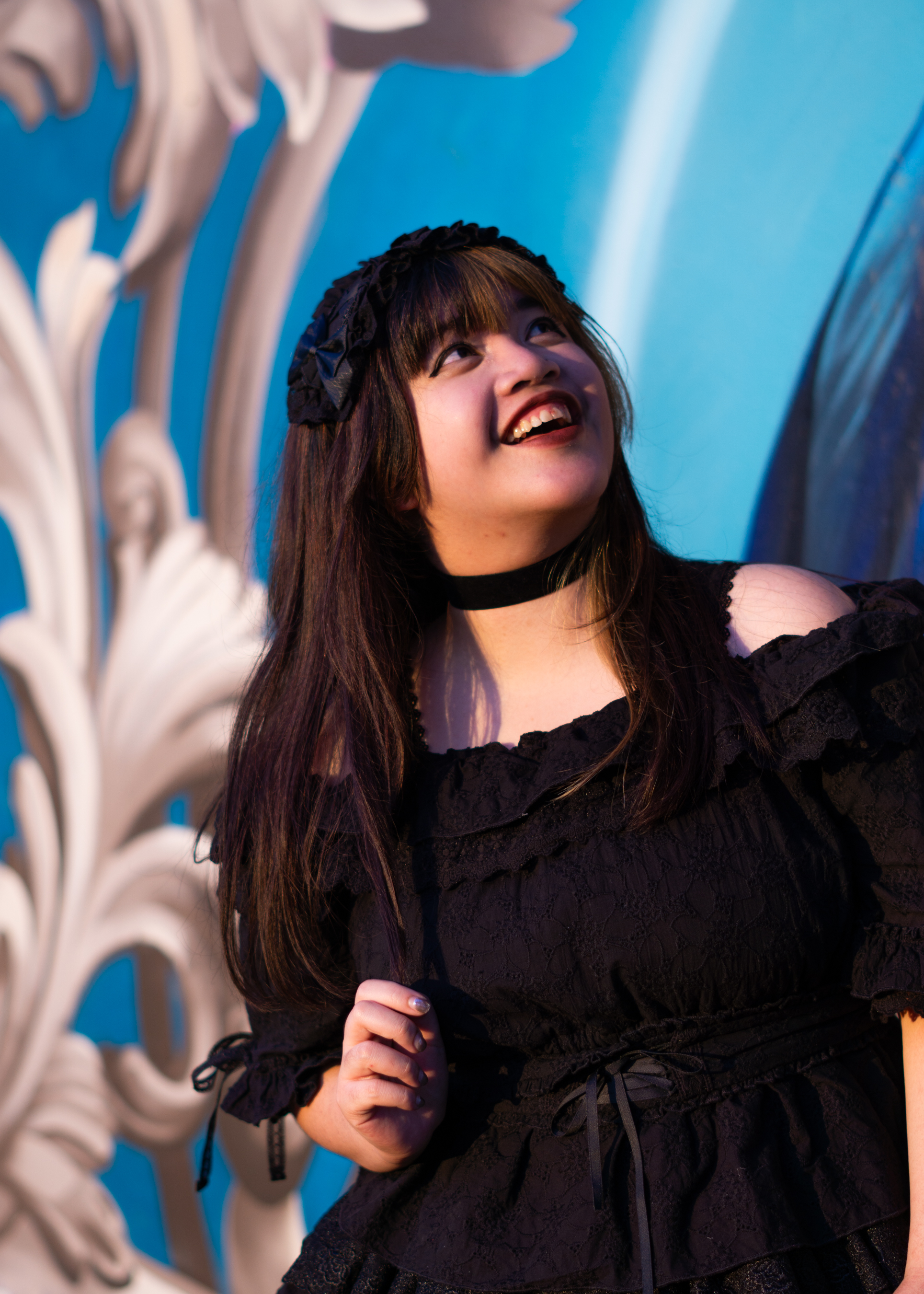 A fem-presenting person in a frilly black dress standing in front of an illustrated wall, blue with white fleurs-de-lys. They're looking up with a smile.