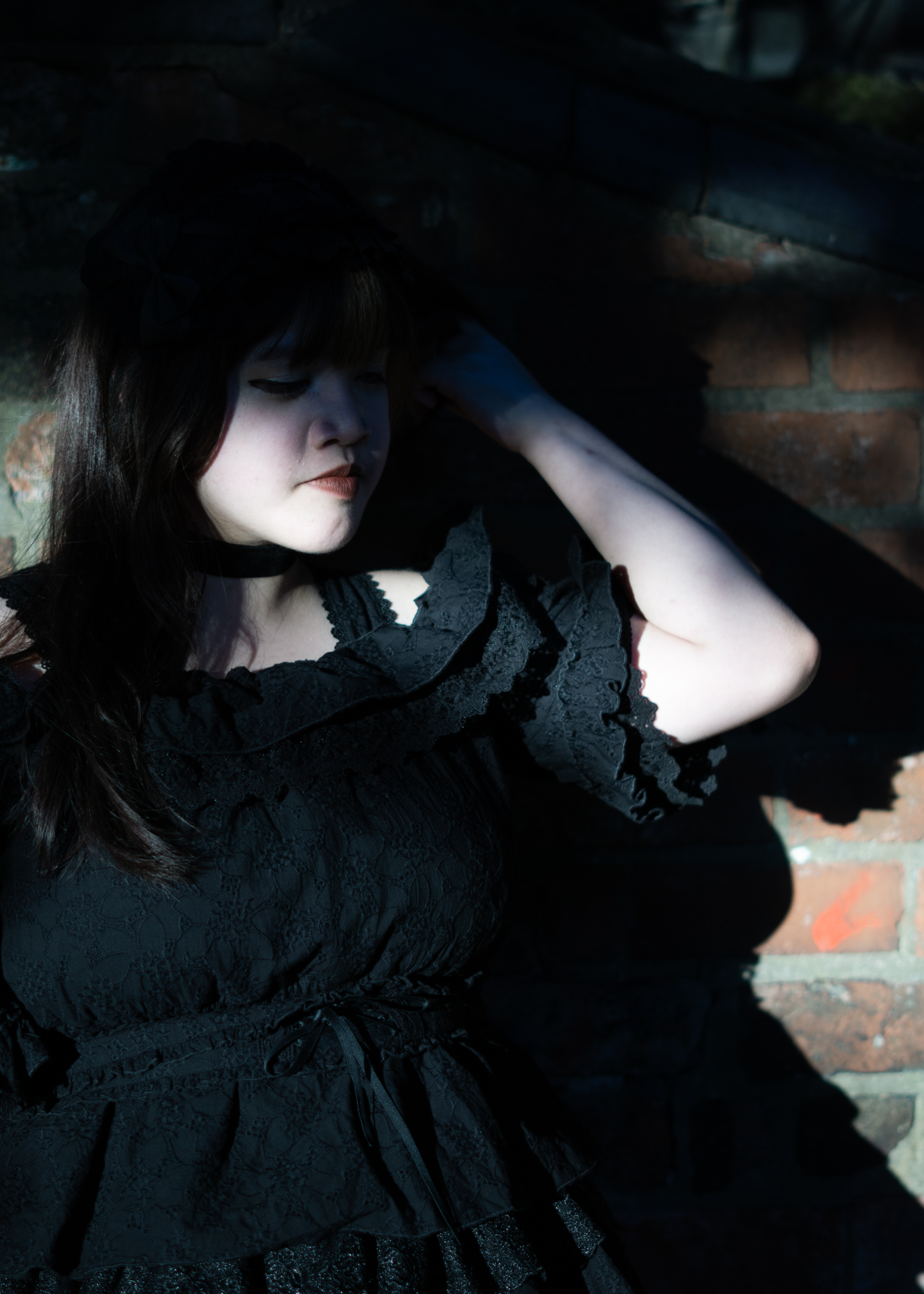 A fem-presenting person in a frilly black dress standing in front of a brick wall, covered mostly in shadow with a streak of sunlight illuminating a stretch of their face. Their face is contemplative and they hold a hand to their chin. There's bright green foliage in the background.