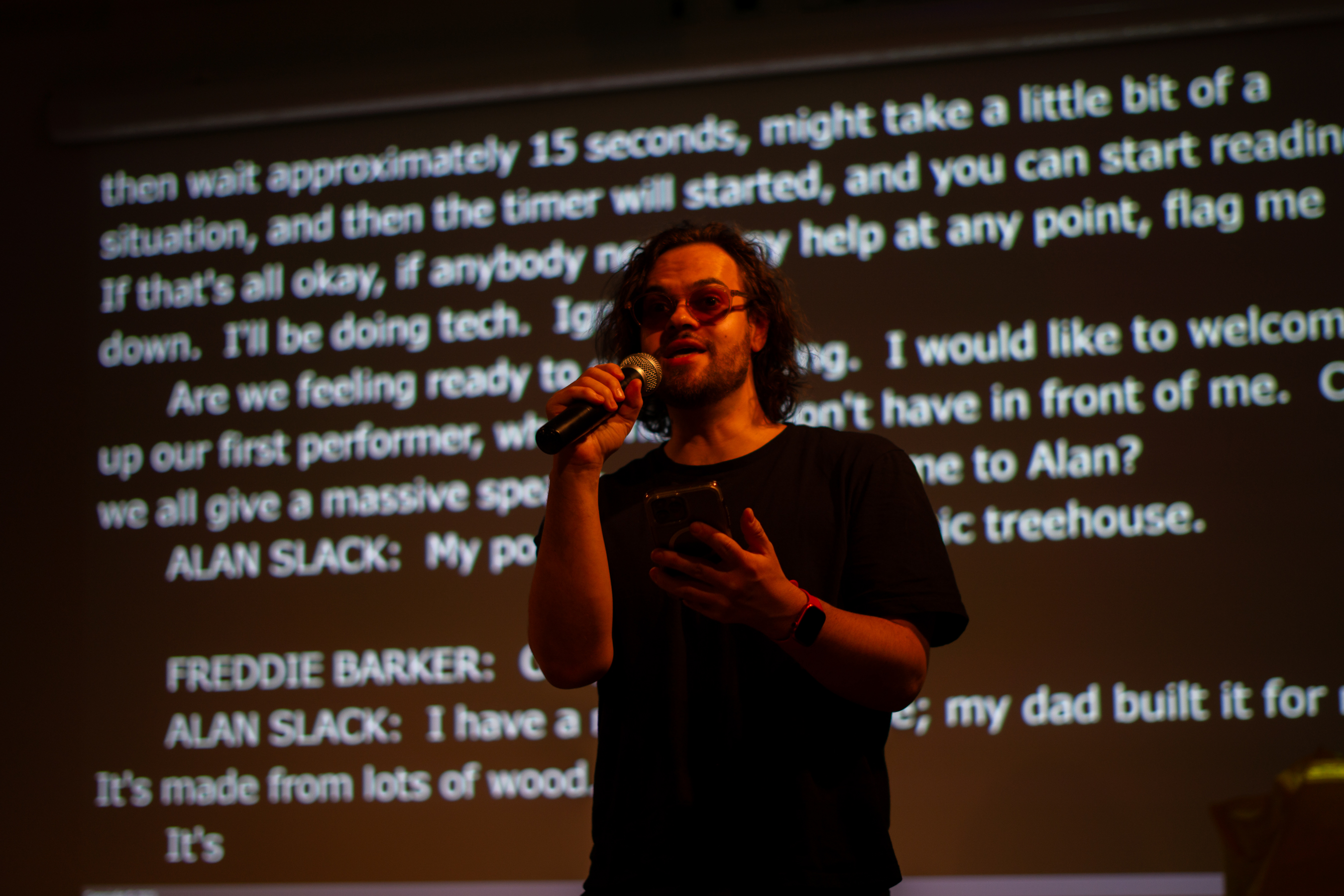 A performer at the Speak Volumes! 2025 poetry slam competition in orange lighting with their poetry projected onto the wall behind them.