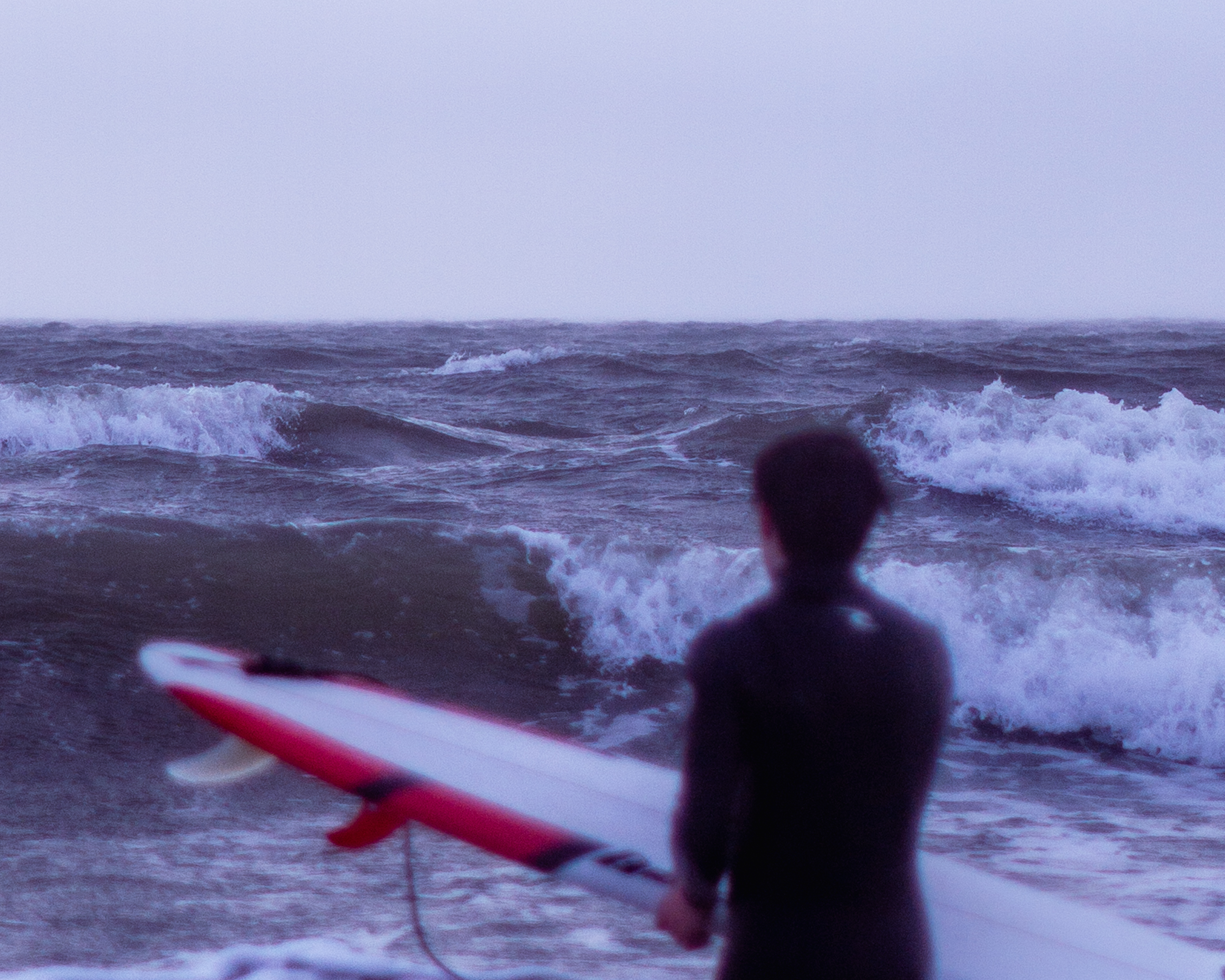 A blurred man in a wetsuit clutching a surfboard blurred in the foreground, waves in focus in the background.