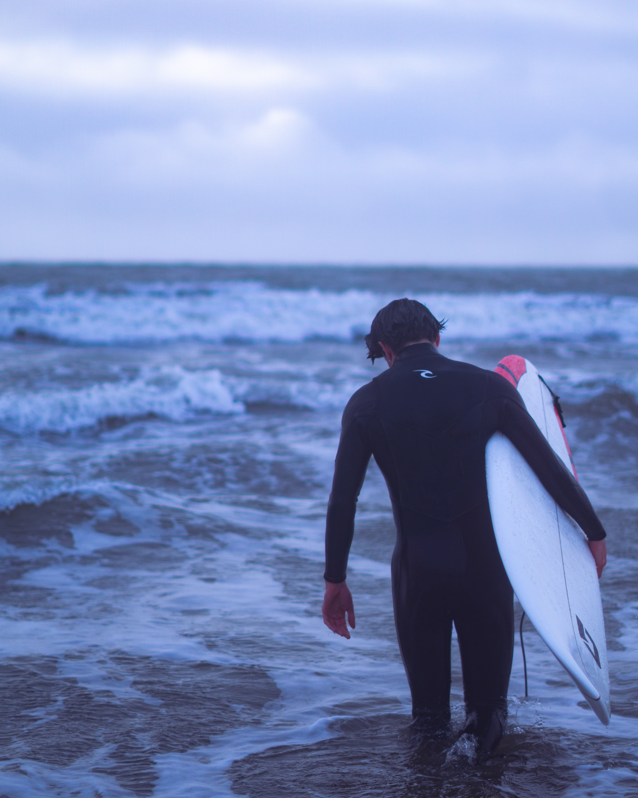 A man in a wetsuit clutching a surfboard and looking down at the gentle waves at his feet.